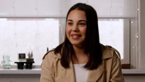 Een foto van Semma in haar woonkamer met op de achtergrond ramen waar het rolgordijn een stuk naar beneden zit en waar witte snoeren met brandende lampjes naar beneden hangen tot aan de vensterbank. 