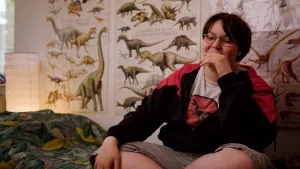 Een foto van Li Platt die op bed zit en een t-shirt met een dinosaurus erop aanheeft. Op de achtergrond hangen posters van getekende dinosaurussen. 