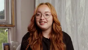 Een foto van Zoë Wijbenga in haar woonkamer met op de achtergrond een muur en een raamkozijn waarop een stukje van een foto zichtbaar is.