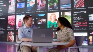 Een foto van presentator Ciana en gast Martijn aan een tafel. Op de tafel staat een laptop. Achter hen is een groot scherm met fimpjes.....