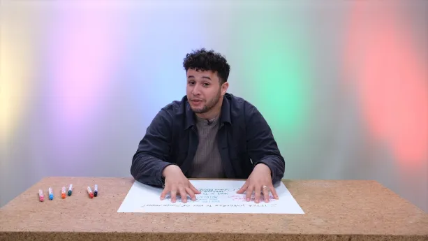 Een foto van Meneer Khalil die achter een tafel zit met zijn handen op een groot vel. Naast het vel liggen zes stiften.