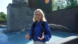 Een foto van presentator Janouk die aan het presenteren is naast het water van een monument. Op de achtergrond is er een beeld te zien.