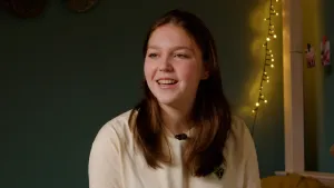 Een foto van een jongere, met op de achtergrond een muur en wat kerstlampjes. Ze praat tegen iemand naast de camera.