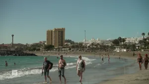 Een foto van een strand op de Canarische Eilanden. Op de voorgrond lopen er twee mannen en een vrouw met de voeten in ondiep water.  Achter hun lopen er meer mensen en zwemmen er een aantal in het water. Op de achtergrond is de stad te zien.