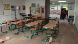 Een foto van een leeg klaslokaal. Op de achtergrond lopen er een aantal leerlingen in de gang weg van het lokaal.