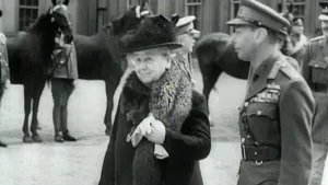 Een foto van koningin Wilhelmina met een handschoen in haar hand. Ze staat naast een soldaat en op de achtergrond staan er paarden met soldaten ernaast.