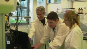 Een foto van drie laboratorium medewerkers die in een laboratorium achter een computer staan. Een van de medewerkers wijst naar iets op het beeldscherm.
