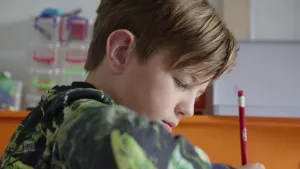 Een foto van een jongen van de zijkant. De jongen heeft een potlood in zijn hand en zit aan tafel te tekenen.