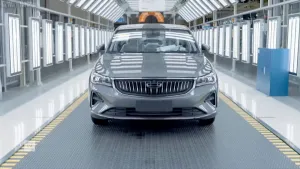 Een foto van een auto die op de lopende band staat in een fabriek, met de neus naar de camera toe. De auto staat in een gang vol met lichten.