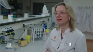 Een foto van een medische specialist die aan het woord is in een laboratorium. Ze staat naast een tafel waar veel laboratorium spullen op liggen.