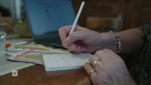 Een foto van de handen van een oudere vrouw met een potlood in haar hand. Zij heeft wat in een aantekeningen boekje geschreven. Er liggen folders onder het boekje en er staat een laptop naast.