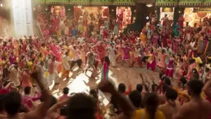 Een foto van een filmset in India waar mensen in een cirkel aan het dansen en zingen zijn. De mensen hebben opvallend gekleurde, wijde kleding aan.