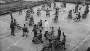 Een zwart-wit foto van een groep mensen in een rolstoel op een basketbalveld in de buitenlucht. De  meeste mensen doen rekoefeningen en drie mensen zijn aan het gewichtheffen met kleine gewichten.