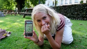 Een foto van presentator Janouk die geknield op een grasveld zit en leunt op haar ellebogen. Ze toont haar telefoon met op het scherm een foto van het grasveld. Op de achtergrond zijn twee picknicktafels zichtbaar.
