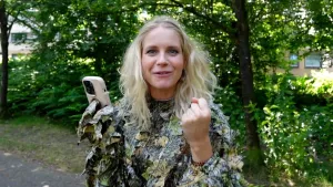 Een foto van presentatrice Janouk die een opvallende blouse aanheeft waarop blaadjes van stof genaaid zijn. Ze heeft een mobiel in haar rechterhand en balt haar linkerhand in een vuist. Achter Janouk zijn diverse bomen zichtbaar.