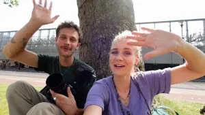 Een foto van presentatoren Janouk en Bram die tegen een boom zitten en zwaaien naar de camera. Bram heeft zelf ook een fotocamera in zijn linkerkand.