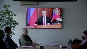 Een foto van een kamer waarin enkele mensen aan een grote tafel zitten. Aan de muur hangt een groot beeldscherm. Daarop zie je de Russische dictator Poetin. De mensen in de kamer luisteren naar hem.