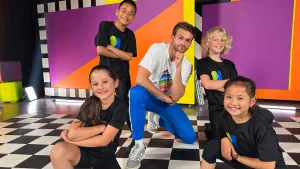 Een foto van danscoach Olivier Feiken die in het midden hurkt op een zwart-wit geblokte dansvloer. Op de voorgrond van de foto staan twee meisjes en naast de danscoach staan twee jongens. De kinderen dragen een t-shirt met hetzelfde logo zonder tekst. 