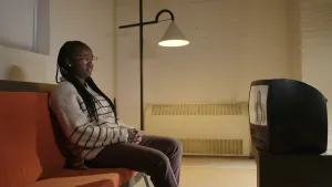 Een foto van presentator Tirsa die op een bank zit achter een kleine televisie. Ze kijkt naar een aflevering van het Klokhuis waar ze zelf aan het woord is.