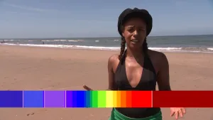 Een foto van een presentator in zwemkleding aan de kust met op de achtergrond de zee. Onderaan is een balk met daarop een kleurverloop te zien van blauw naar rood. De balk met kleuren wordt gebruikt bij het vertellen over de hoeveelheid zonnestraling.