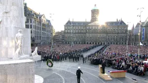 Een foto van de kranslegging op de dodenherdenking. Er is een krans gelegd en er staat een groep mensen er tegenover met publiek aan beide kanten. Links op de voorgrond is het Nationaal Monument te zien.