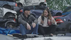 Een foto van presentator Pascal die praat met Pieter Koolwijk terwijl ze op een auto wrak zitten. Op de achtergrond zijn er meer auto wrakken opgestapeld.