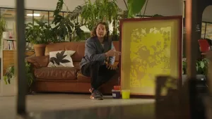 Een foto van presentator Pascal die op een bank zit en over een groen kleurige ingelijste zeefdruk praat die naast hem staat.