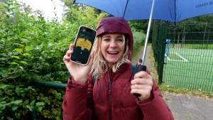 Een foto van presentator Janouk met een rode regenjas aan. In haar linkerhand is een paraplu zichtbaar, in haar rechterhand een mobiele telefoon, waarvan ze het scherm naar de camera richt. De tekst op het scherm is niet leesbaar.