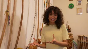 Een foto van Serah die naast een muur staat waar Capoeira muziek instrumenten hangen. Achter haar staan er drums op de grond en hangen er foto lijstjes aan de muur.