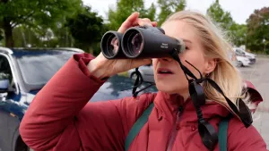 Een foto van presentator Janouk in een straat die met een verrekijker naar een punt kijkt achter de camera.