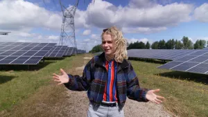 Een foto van presentator Janouk die buiten op een pad loopt tussen velden met zonnepanelen in.