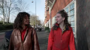 Een foto van presentator Serah die met een vrouw praat terwijl ze langs huizen over de stoep lopen.