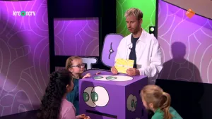Een foto van de presentator bij een grote doos met ogen erop en een gleuf aan de voorkant. Rond de doos staan drie kinderen die naar de doos en de presentator kijken.