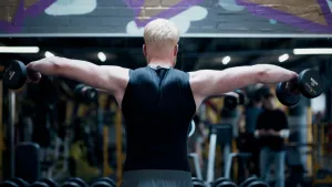 Een foto van een man die met gestrekte armen twee halters vasthoudt en met zijn rug naar de camera toe staat.