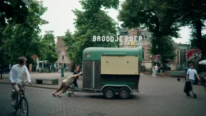 Een foto van een omgebouwde paarden aanhanger die door twee mensen op de plaats wordt geduwd op straat. Op het dak staat de tekst broodje poep. Op de voorgrond is er een fietser zichtbaar die ernaar kijkt.