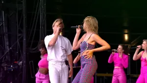 Een foto van een zanger en een zangeres die zingen met een microfoon in hun hand. Op de achtergrond zijn één danseres en drie zingende achtergrondzangeressen zichtbaar.