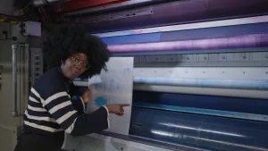 Een foto van een presentator die staat voor een drukpers in een drukkerij en een nog te drukken vel papier vasthoudt.