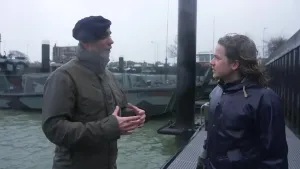 Een foto van een marinier in gesprek met presentator Pascal op de kade in een haven. Achter hen ligt een militair voertuig in het water dat zowel op land als op zee kan.