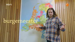 Een foto van presentator Pascal bij een grote kaart van Nederland. Op de kaart hebben alle gemeenten verschillende kleuren. Over de foto staat de tekst "burgemeester".