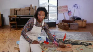 Een foto van presentator Astrid die naar Berlijn wijst op een kaart van Duitsland van na de tweede wereldoorlog. De kaart ligt op tafel en geeft de gebieden aan waar Westerse landen de macht hebben, aangegeven door vlaggen die bovenop de kaart staan.