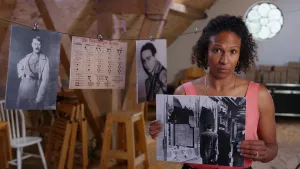 Een foto van een presentator die een zwart wit foto vasthoudt met daarop drie mensen die voor een gebouw staan met kapotte ramen. Op de achtergrond hangt er een zwart wit foto van Adolf Hitler aan een lijn.