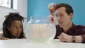 Een foto van presentator Eva en hersenwetenschapper Marsh die staan aan een tafel bij een vissenkom. In de vissenkom zitten een stel hersenen van een mens in vloeistof. Op de achtergrond is een anatomisch model van een mens zichtbaar.