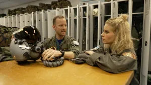 Een foto van een piloot en presentatrice Janouk die allebei legerkleding dragen. Ze staan bij een hoge tafel met een pilotenhelm erop.