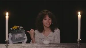 Een foto van presentator Serah die achter een tafel zit en met haar duim naar een accu met daarop een speelgoed auto wijst.  Voor haar staat een kommetje met yoghurt en ze zit tussen twee aangestoken kaarsen in.