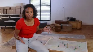 Een presentator zit op een tafel waar twee landkaarten op liggen. Bovenop verschillende landen staan er plastic speelgoed soldaatjes met twee verschillende kleuren.