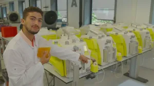 Een foto van een man die in een laboratorium staat en naar een bloed scheidingsapparaat wijst. Op de tafel staan er nog meer bloed scheidingsapparaten. 
