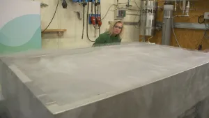 Een foto van presentator Janouk die achter een bak met koud water staat. Er komt damp af van het water en op de achtergrond zitten er apparaten, buizen en kabels aan de muur.