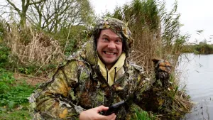 Een foto van een presentator in onopvallende kleding die camouflagekleding genoemd wordt in het gras vlak aan de waterkant. In zijn rechterhand heeft hij een mobiele telefoon vast.  