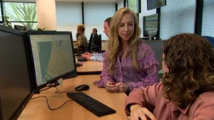 Een foto van presentator Pascal en Bianca van de ANWB die achter een bureau zitten op het kantoor van de ANWB. Links van hen is een beeldscherm te zien met daarop een van een gedeelte van Nederland en, op de foto niet leesbare, verkeersinformatie.