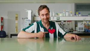 Een foto van een presentator die aan een tafel zit in een laboratorium. Hij wijst met z'n wijsvinger naar een getekend hart op zijn borst.